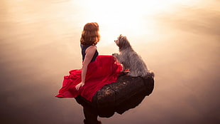 woman in black and red dress sitting beside long-coated gray dog on rock in middle of water
