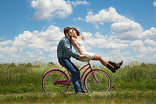 man on blue checkered flannel kissing by a girl wearing white spaghetti strap dress riding on red bicycle during day time