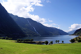 green open field, body of water, and mountains under blue skies and white clodus