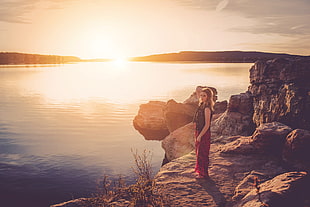 woman in black and red dress standing on a rock near body of water during golden hour