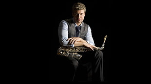 man wearing grey vest and grey dress shirt sitting while holding his brass-colored saxophone