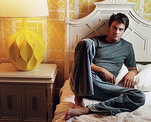 man wearing gray shirt and jeans sitting on white bed near yellow table lamp