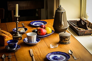 fruits on blue and white ceramic plate beside gray teapot and gray metal candle holder with white taper candle on brown wooden table