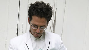 man in white blazer and eyeglasses standing near white wall