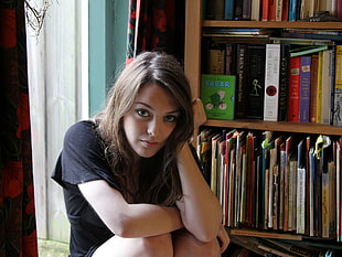 woman wearing black t-shirt sitting near brown wooden book shelf at daytime