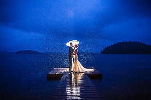 photo of couple standing on wooden dock near body of water while holding umbrella
