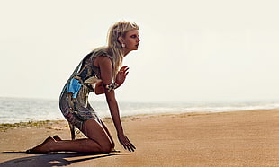 blond haired woman in gray and white sleeveless dress kneeling on beach sand