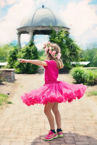toddler girl wearing pink tutu dress with pink-and-green Sketchers running shoes standing on brown concrete brick pathway near white dome gazebo during daytime