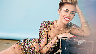 Miley Cyrus wearing brown glittered long-sleeved dress lies near black cajon box