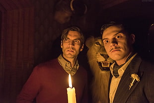 man in suit jacket holding candle standing beside man in maroon shirt and lion
