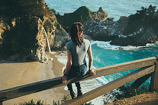 sitting woman wearing blue jeans and gray shirt on wooden railings near sea