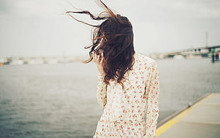 woman wearing white and pink floral long-sleeved shirt covered her face with her hair beside body of water during daytime