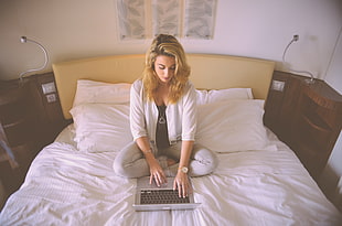 woman in white open cardigan sitting on white bedspread holding MacBook Pro