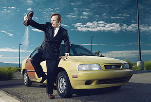 man in black suit jacket and dress pants holding brown loafers standing beside yellow car under blue sky during day time