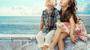 boy and girl sitting on brown concrete seaside seat during daytime HD wallpaper