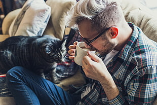 Man wearing black , white and red plaid dress shirt drinking coffee while sitting beside brown tabby cat