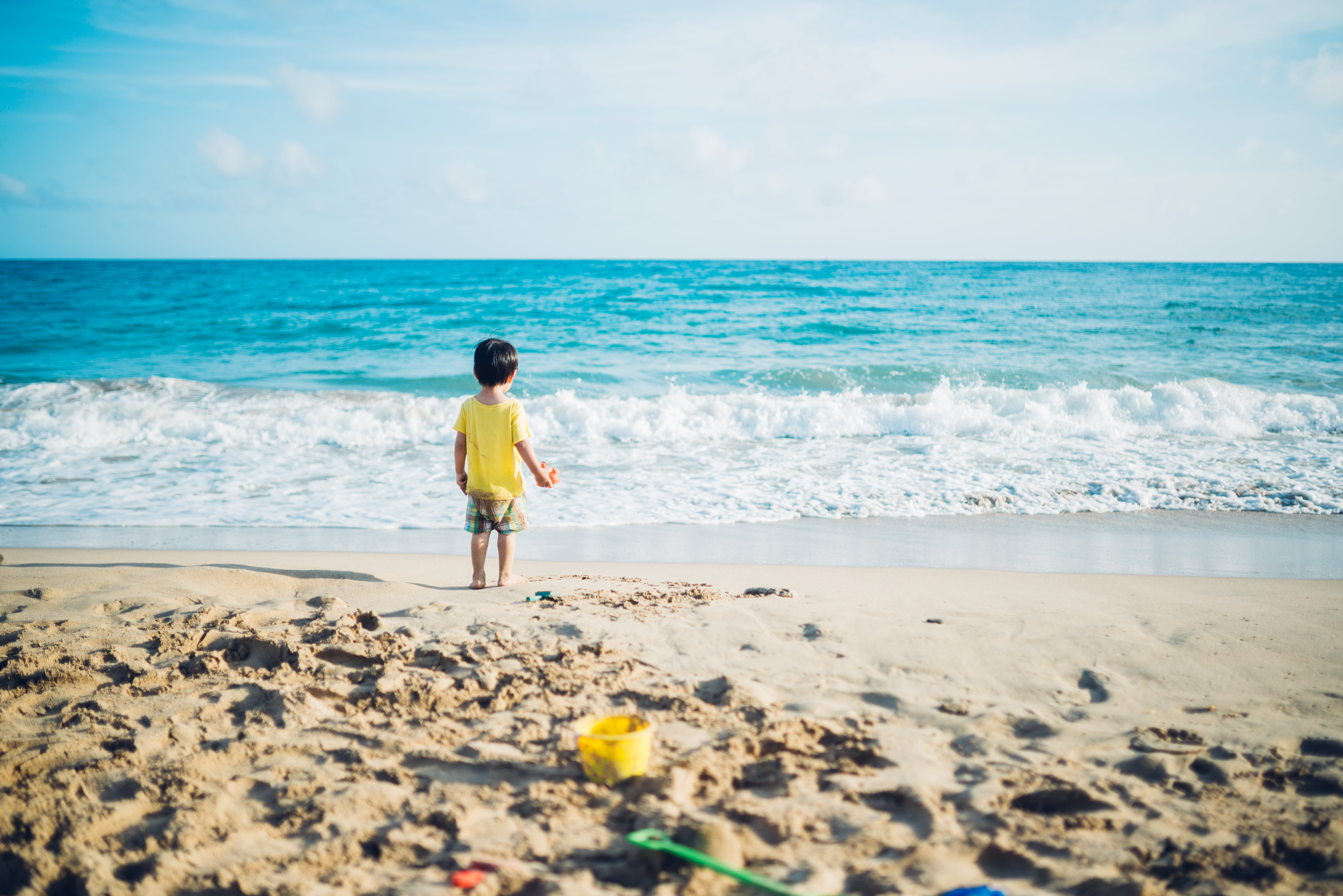 toddler in yellow shirt and gray shorts standing at seashore near blue sea during daytime