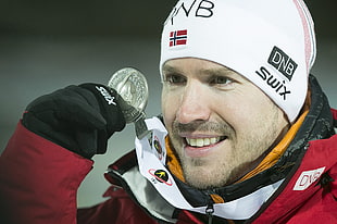 man in red and black jacket wearing white swix beanie hat holding silver medal