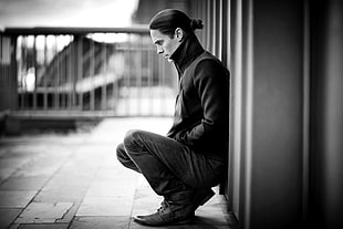 grayscale photo of man wearing black jacket leaning on wall