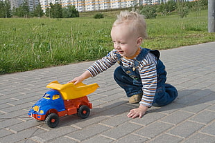 boy in blue denim overalls, white-and-brown striped long-sleeved shirt playing blue-yellow-and-red plastic dump truck on concrete pathway during daytime