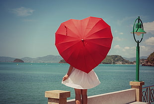 woman wearing white dress holding red heart-shape umbrella standing in front of sea during daytime