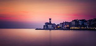 calm body of water with lighted buildings under pink and orange sky, piran