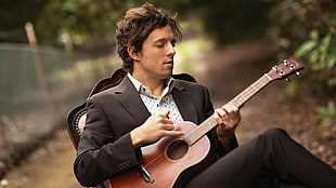 man in black suit playing brown ukulele sitting on white padded chair