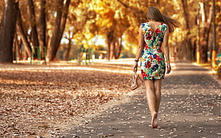 woman wearing floral dress walking in gray pavement road in between brown trees