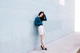 woman in white skirt and teal long sleeve top standing on concrete pavement leaning on teal painted wall