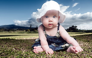 baby wearing blue floral dress and white knit bucket hat sitting on green grass field