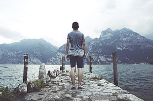 man in grey t-shirt standing grey concrete dock near mountain during daytime