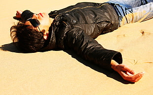 man wearing black aviator sunglasses and leather jacket lying on the sand