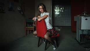woman wearing white and black cap-sleeved shirt and red, black, and white plaid skirt sitting on red padded chair near white painted wall in room HD wallpaper