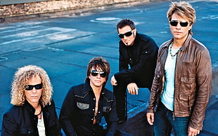 four men wearing black sunglasses with brown and black jackets standing near road photo taken during daytime