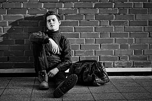 grayscale photography of man sitting and leaning on brick wall with duffle bag