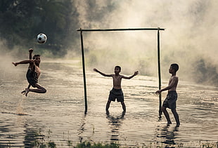 three boys playing soccer on body of water