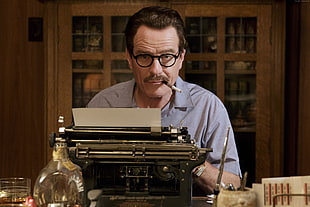 man with gray collared shirt and black framed eyeglasses sits in front of black and gray typewriter