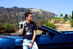 man in blue t shirt sitting side of car door over looking Hollywood hill signage at day time