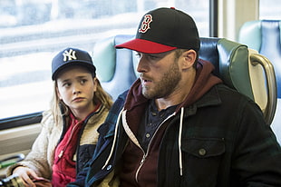 man wearing black Boston Red Sox fitted cap and black jacket beside girl in white and black jacket inside train