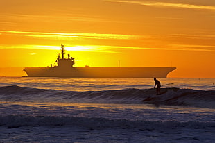 silhouette of person board surfing on body of water with ship during sunset