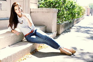 woman wearing white dress shirt and blue denim skinny jeans sitting on brown concrete stair near green plants during daytime