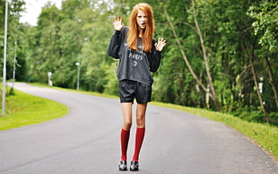 woman wearing gray and black long-sleeved shirt and black shorts outfit with red high socks and black leather shoes standing between gray asphalt road