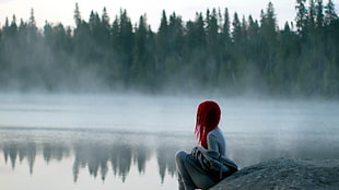 depth of field photography of woman sitting near body of water surrounded by trees