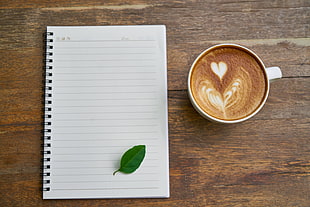 photo of white ceramic coffee cup filled with cup near white ruled notebook on brown wooden board