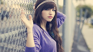 woman wearing purple long-sleeve shirt leaning on grey link fence on selective focus photography