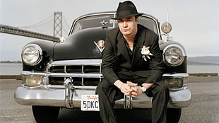 man in black suit sitting on classic black car over looking suspension bridge