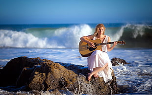selective focus photography of woman in white dress playing dreadnought brown acoustic guitar while sitting on rock against water wave