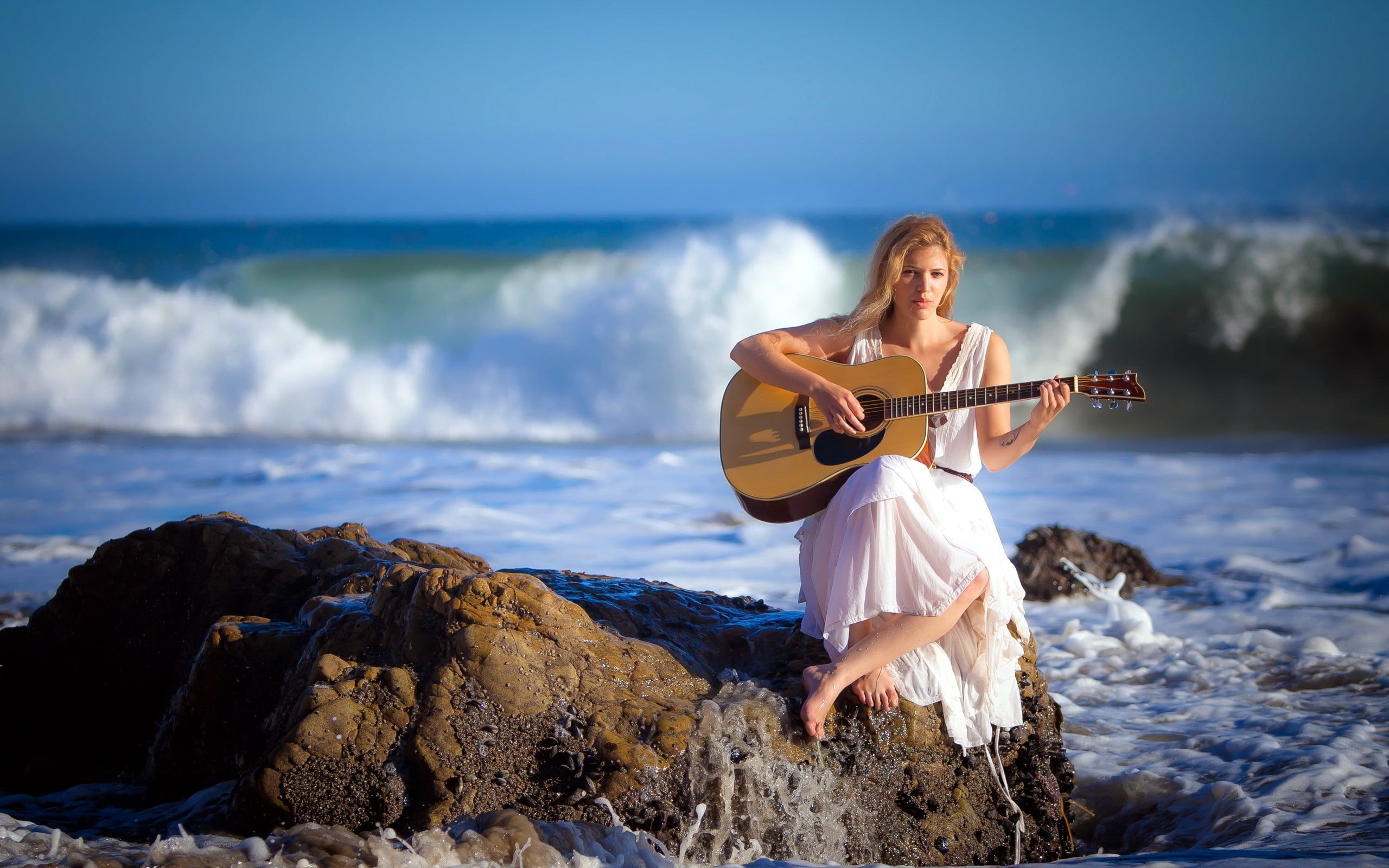 selective focus photography of woman in white dress playing dreadnought brown acoustic guitar while sitting on rock against water wave