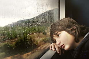 boy wearing black zip-up hoodie staring at glass window with water droplets