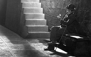 gray scale photo of Charlie Chaplin sitting on bench near stairs
